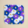 A square fabric cloth with rounded corners, featuring a colourful floral pattern of purple, blue, and white flowers with yellow centers and green leaves, finished with a white stitched border, shown flat on a beige background.