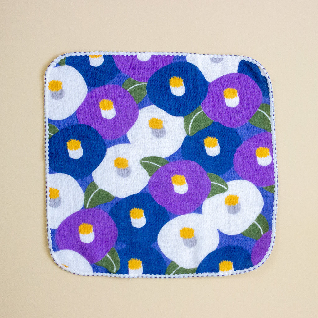 A square fabric cloth with rounded corners, featuring a colourful floral pattern of purple, blue, and white flowers with yellow centers and green leaves, finished with a white stitched border, shown flat on a beige background.