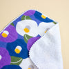A close-up of a folded fabric item, showing a colourful floral print with purple, blue, white, green, and yellow flowers, alongside a soft white fleece lining.