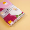 A folded Japanese handkerchief with a colourful floral pattern, wrapped in a paper band that reads 'HAIKARA HANDKERCHIEF made in JAPAN', set against a solid pale yellow background.