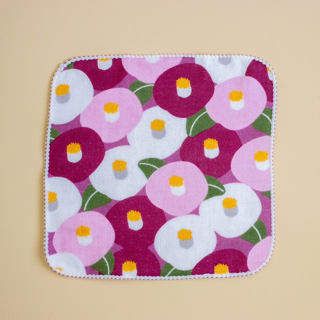 A square fabric item with rounded corners, featuring a colourful floral pattern of pink, white, and magenta flowers with green leaves, finished with a white scalloped edge, displayed flat on a beige background.