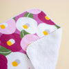 A close-up of a folded fabric cloth with a floral print featuring pink, white, green, and yellow flowers, showing the white textured reverse side.