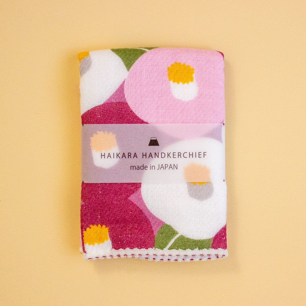 A folded Japanese handkerchief with a colourful floral pattern, wrapped in a paper band that reads 'HAIKARA HANDKERCHIEF made in JAPAN', set against a solid pale yellow background.