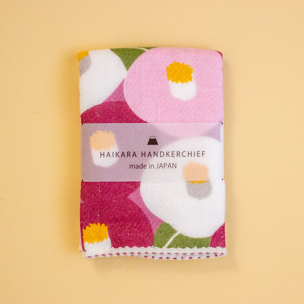 A folded Japanese handkerchief with a colourful floral pattern, wrapped in a paper band that reads 'HAIKARA HANDKERCHIEF made in JAPAN', set against a solid pale yellow background.