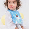 A baby wearing a light blue bib with a cat design and white long-sleeve shirt, photographed against a soft white background with yellow balloons.