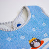 Close-up of a baby bib made of textured blue fabric with a grey and white striped neckline, small white motifs, and a cartoon cat wearing a yellow hat.
