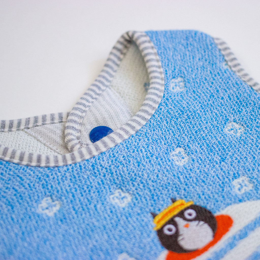 Close-up of a baby bib made of textured blue fabric with a grey and white striped neckline, small white motifs, and a cartoon cat wearing a yellow hat.