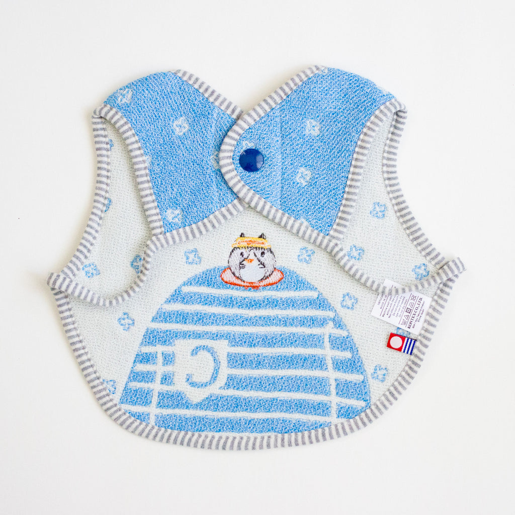 A baby bib with a light blue background and white cat design, featuring grey striped trim and a snap closure, laid flat on a white background.
