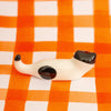 A small glossy ceramic figurine of a cat lying down, featuring an off-white body with black spots, resting on an orange and white checkered background.