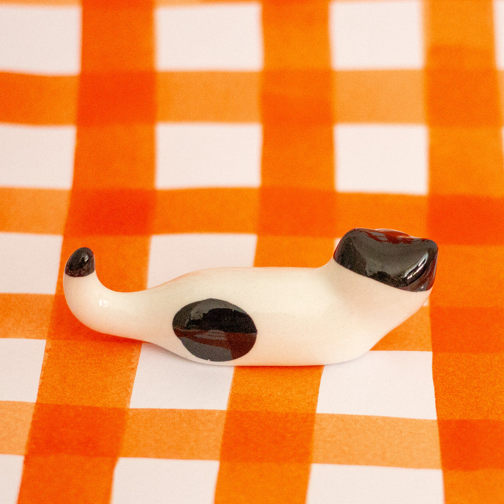 A small glossy ceramic figurine of a cat lying down, featuring an off-white body with black spots, resting on an orange and white checkered background.