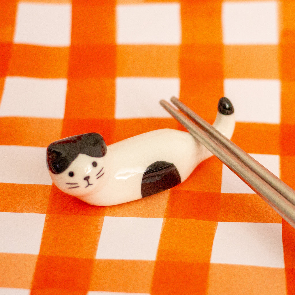 A small glossy cat-shaped chopstick holder, shown lying on an orange-and-white checkered surface with metal chopsticks resting near it.