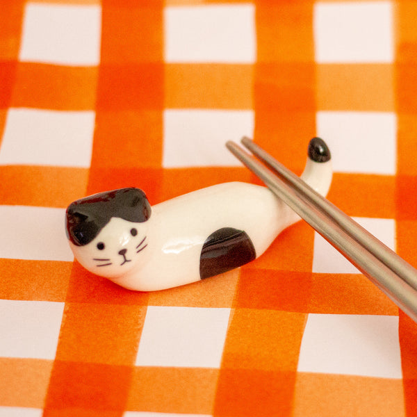 A small glossy cat-shaped chopstick holder, shown lying on an orange-and-white checkered surface with metal chopsticks resting near it.