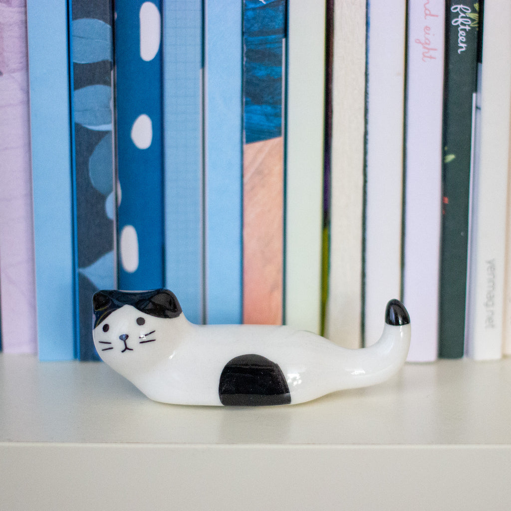 A small glossy ceramic cat figurine in white with black patches, positioned on a white shelf in front of a row of colourful books.