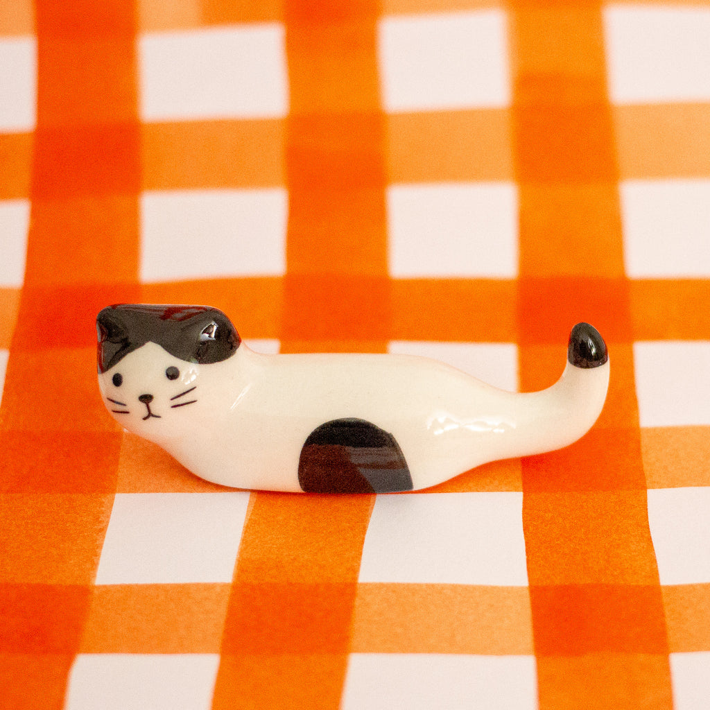A small glossy cat figurine with a white body, black patches, and black-tipped tail and paw tips, posed on an orange-and-white checkered surface.