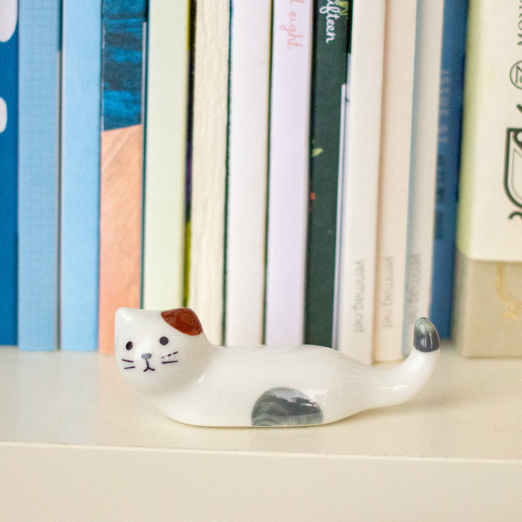 A small white ceramic cat figurine with brown and grey accents, positioned on a light-coloured shelf in front of a row of colourful books.