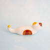 A small white ceramic figurine of a cat lying down, with brown and yellow accents, set against a soft light blue background.
