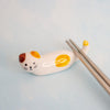 A small white ceramic cat-shaped chopstick rest with yellow and brown patches, holding a pair of metal chopsticks on a pale blue background.