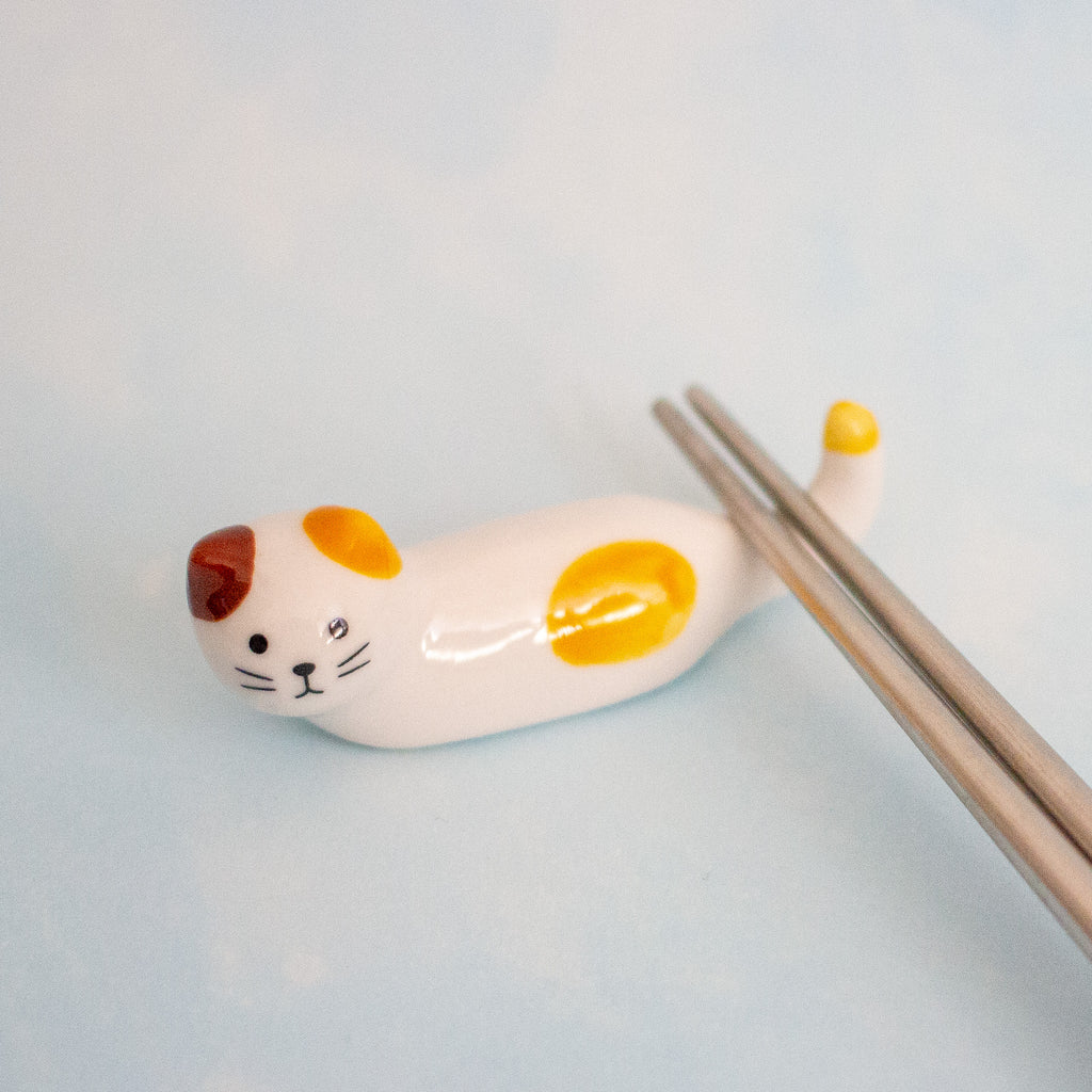 A small white ceramic cat-shaped chopstick rest with yellow and brown patches, holding a pair of metal chopsticks on a pale blue background.