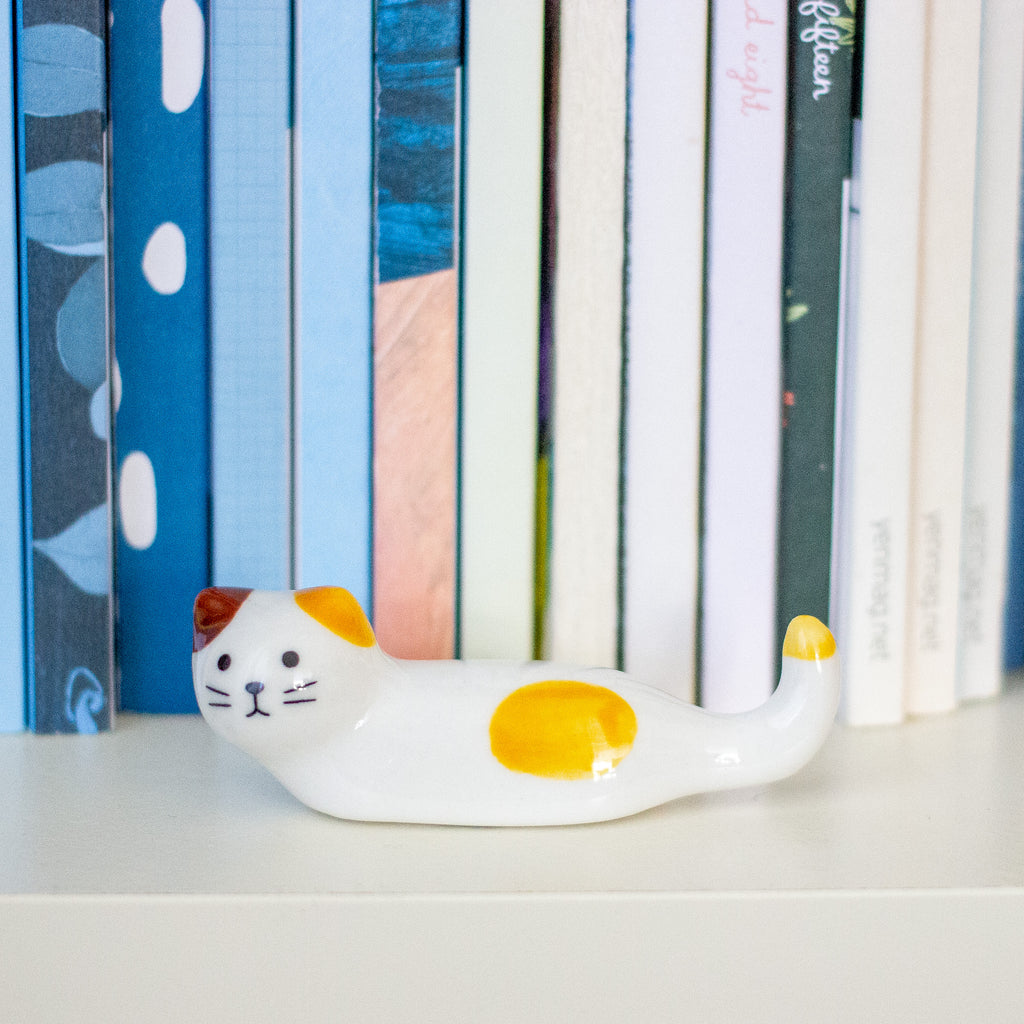 A small ceramic cat figurine in white with yellow and orange patches, placed on a white shelf in front of a row of colourful books.