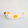 A small white ceramic cat figurine white with yellow patches, brown ears, and black facial details, resting on a light blue background.