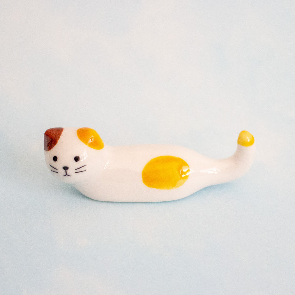 A small white ceramic cat figurine white with yellow patches, brown ears, and black facial details, resting on a light blue background.