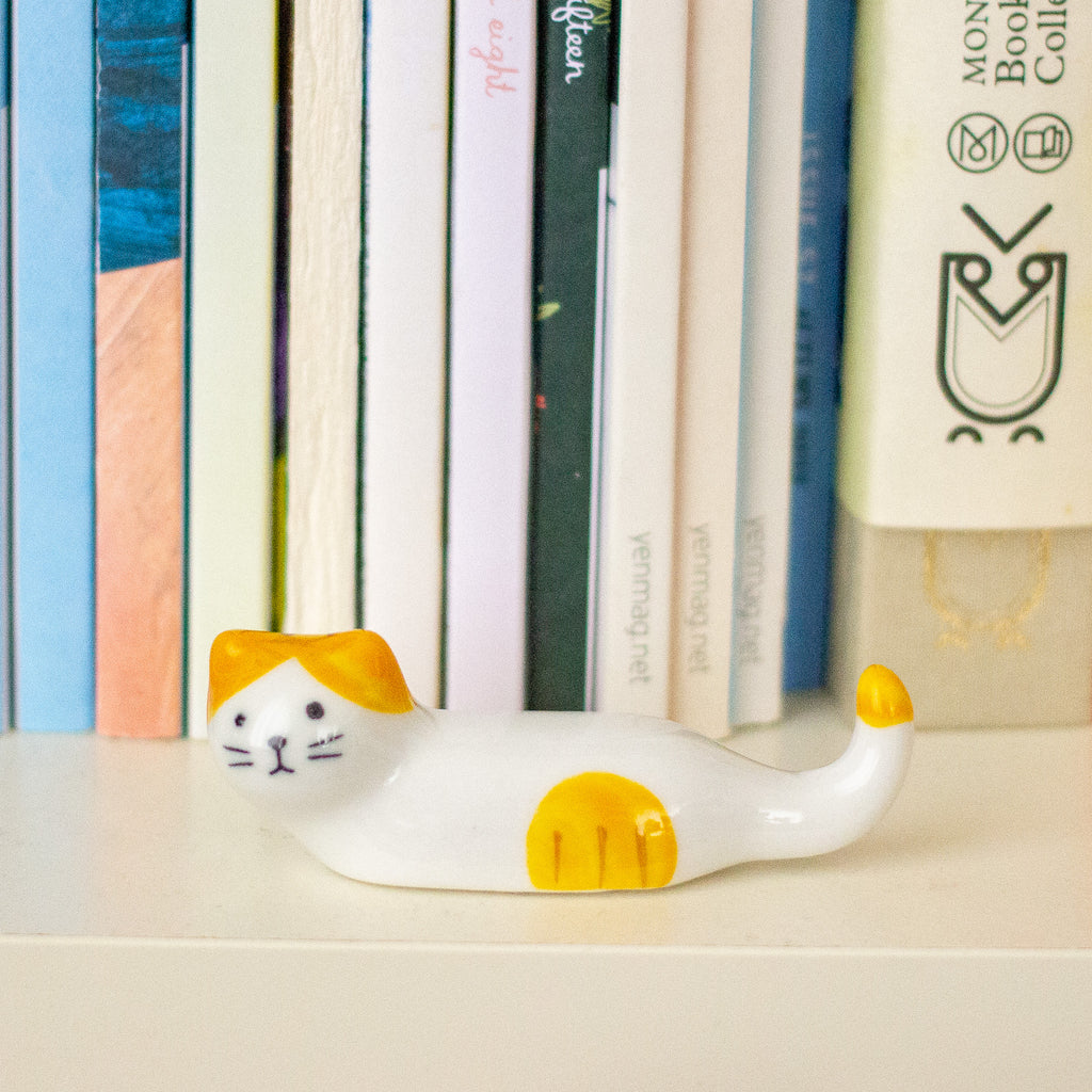 A small ceramic cat figurine in a curled-up resting pose, decorated with a white body and yellow accents on its head, paws, and tail tip, displayed on a shelf in front of a row of colourful books.