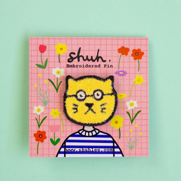 Embroidered pin with a cat face design on a pink grid background with flowers, brand 'shuh.' visible.
