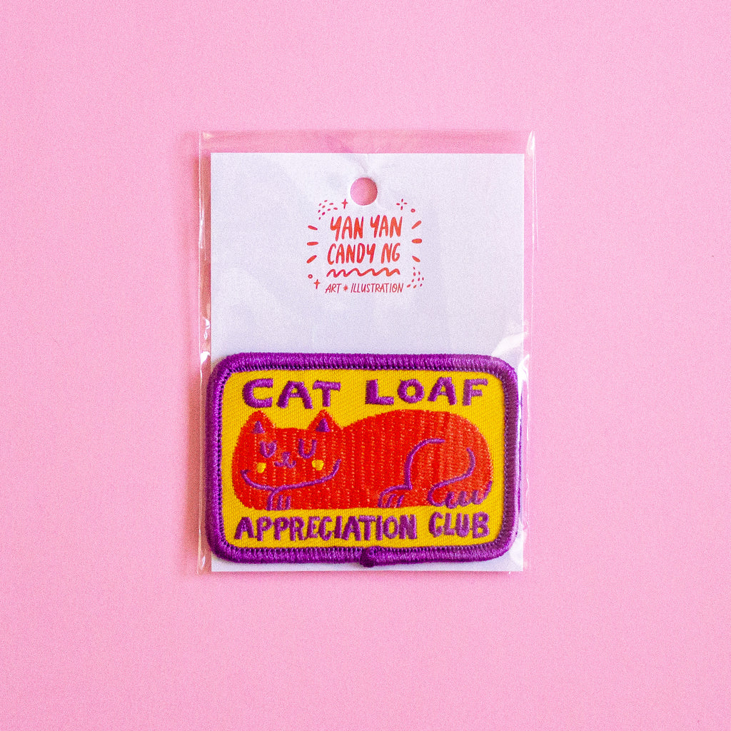 An embroidered patch featuring a sleeping orange cat on a yellow background with purple text reading 'CAT LOAF APPRECIATION CLUB' and a purple border, attached to a white backing card with the brand 'Yan Yan Candy Ng' visible, set against a solid pink background.