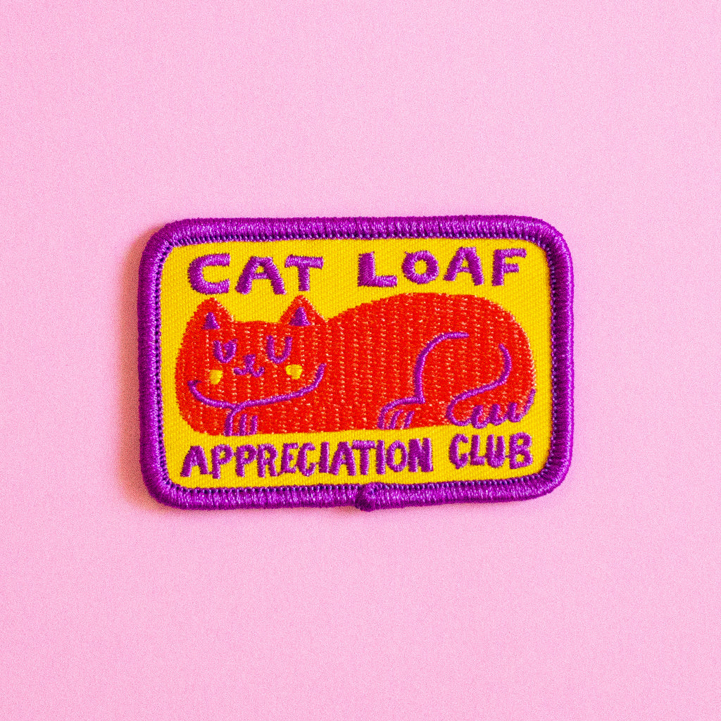 An embroidered patch featuring a red cat on a yellow background with purple text reading 'CAT LOAF APPRECIATION CLUB' and a purple border, placed on a solid pink background.