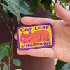 A hand holding a rectangular embroidered patch that reads 'CAT LOAF APPRECIATION CLUB' with a purple border, yellow background, and an orange cat illustration, set against a background of green leaves and a brown wall.