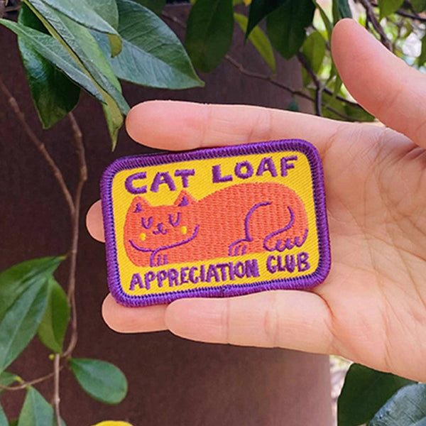 A hand holding a rectangular embroidered patch that reads 'CAT LOAF APPRECIATION CLUB' with a purple border, yellow background, and an orange cat illustration, set against a background of green leaves and a brown wall.