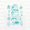 A white kitchen tea towel featuring a teal line art illustration of a stacked cat tower and surrounding potted plants, displayed flat on a white wood plank background.