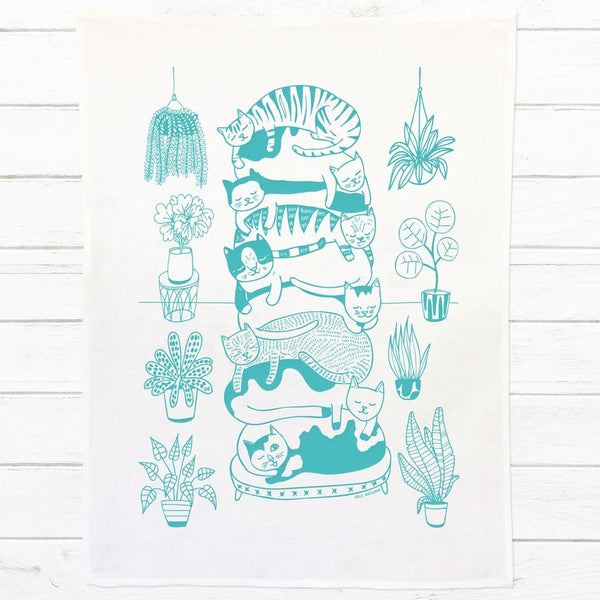 A white kitchen tea towel featuring a teal line art illustration of a stacked cat tower and surrounding potted plants, displayed flat on a white wood plank background.
