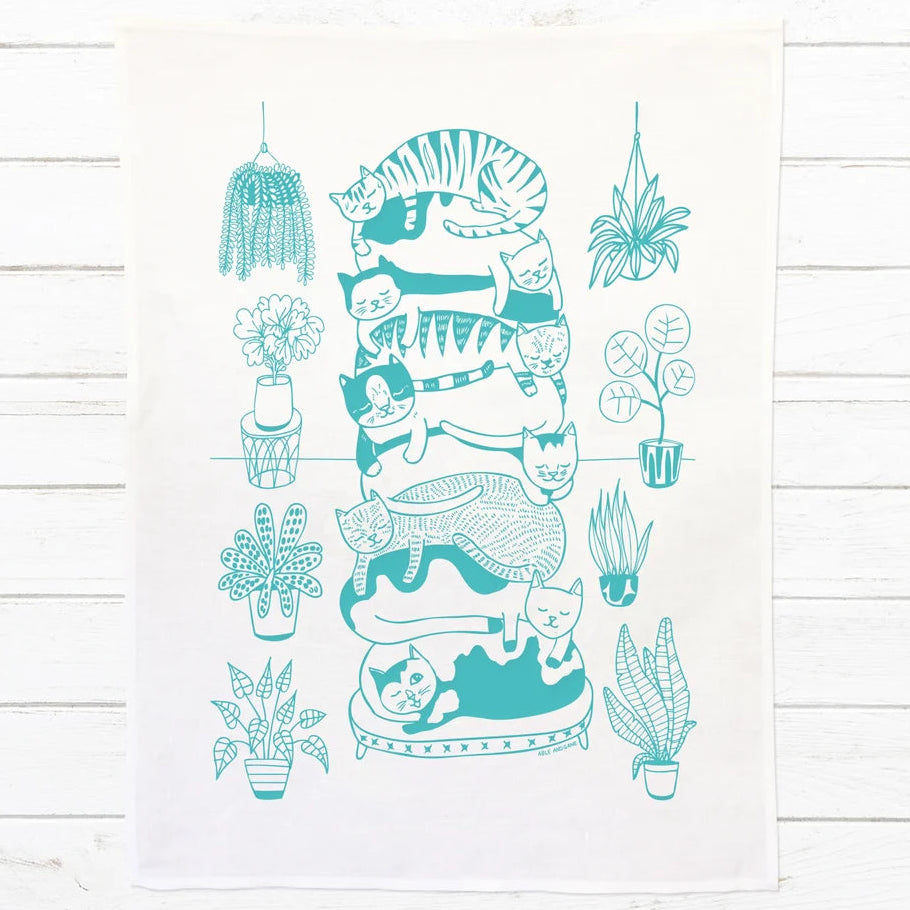 A white kitchen tea towel featuring a teal line art illustration of a stacked cat tower and surrounding potted plants, displayed flat on a white wood plank background.