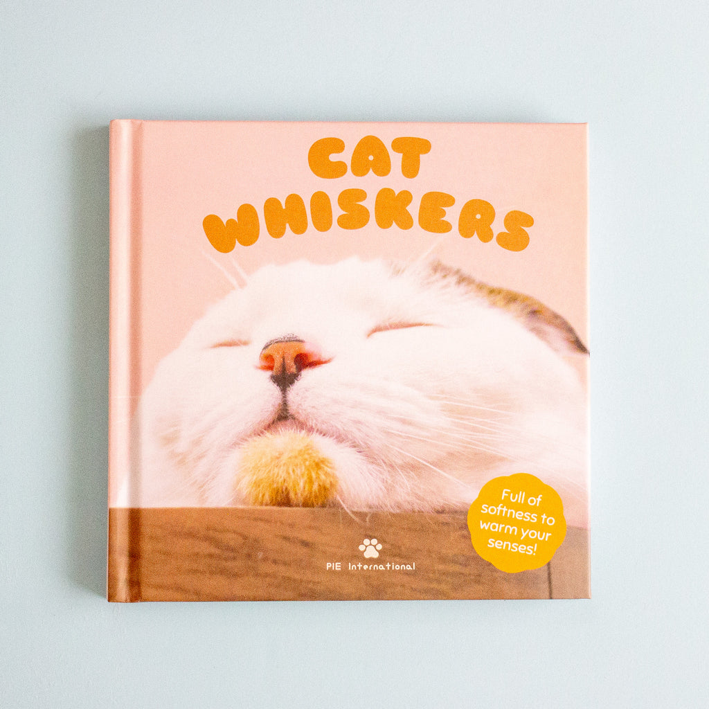 A hardcover book titled 'CAT WHISKERS' with a close-up photo of a sleeping white cat on the cover, photographed against a light blue background.
