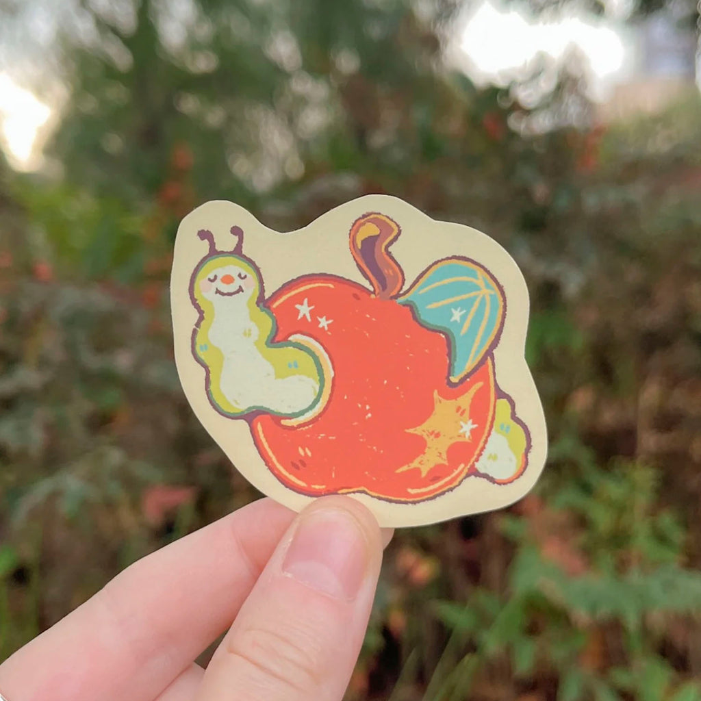 A hand holding a die-cut sticker featuring a whimsical illustration of a red apple with a smiling green worm and a blue winged creature, set against a blurred outdoor background.