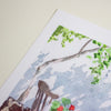 A close-up of a watercolour painting on textured paper, showing green leaves, tree branches, a brown chair, and small red flowers against a pale blue and white background, placed on a light beige surface.