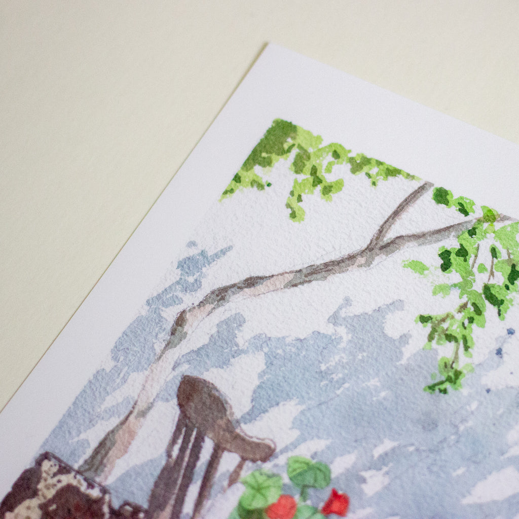 A close-up of a watercolour painting on textured paper, showing green leaves, tree branches, a brown chair, and small red flowers against a pale blue and white background, placed on a light beige surface.