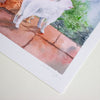 A close-up of a watercolour art print showing a white cat standing on terracotta-coloured paving stones with green foliage in the background. The print has a wide white border and a cursive artist signature near the bottom, photographed on a light neutral surface.
