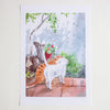 A watercolour art print depicting an orange and white cat standing on a tiled patio next to a potted plant, with a wooden chair and tree in the background. The print is presented on a white paper sheet against a light beige background.