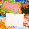A blank white rectangular postcard with a small round blue stamp sits centered on top of a colourful collage of illustrated cat-themed papers and patterned sheets.