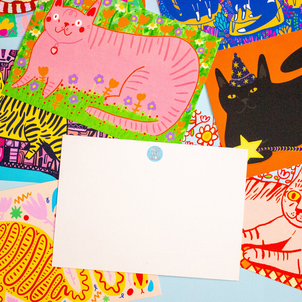 A blank white rectangular postcard with a small round blue stamp sits centered on top of a colourful collage of illustrated cat-themed papers and patterned sheets.