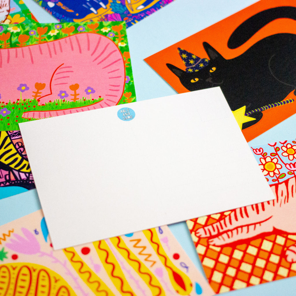 A blank white rectangular postcard lies on top of several colourful illustrated cards featuring a black cat, flowers, stripes, and abstract patterns on a light blue background.