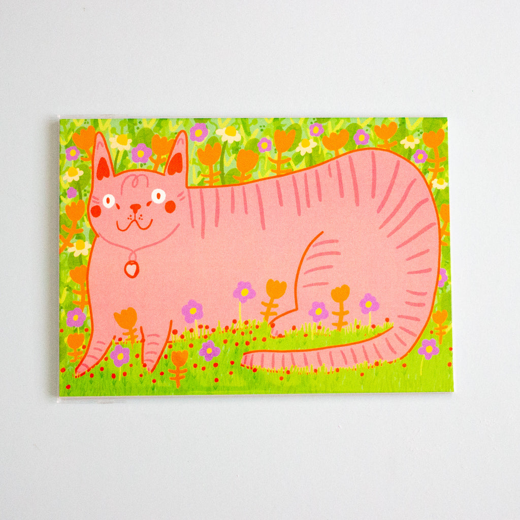 A postcard featuring a whimsical pink cat surrounded by a green field of flowers, shown against a plain white background.