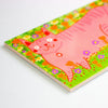 A close-up of a colourful printed postcard featuring a pink cat lying on a green grassy field with small flowers, photographed on a light neutral background.