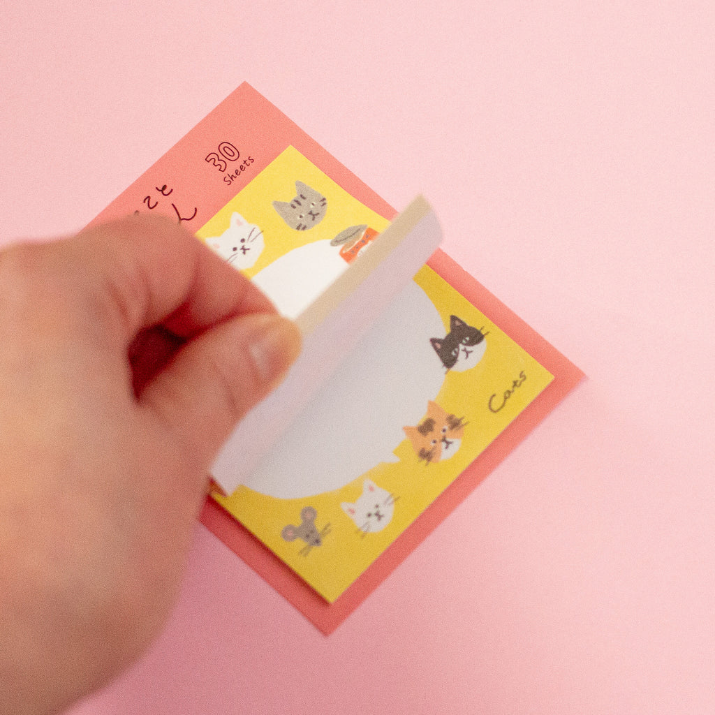 A hand peeling back a yellow sticky note pad with cute cartoon cat illustrations, placed on top of a coral-pink backing sheet against a solid pink background.