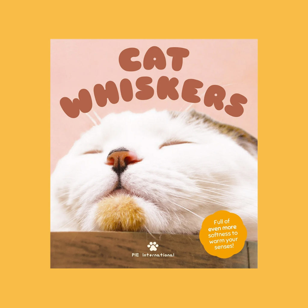 A square book cover titled "CAT WHISKERS" featuring a close-up photo of a sleeping white cat on a wooden surface against a soft pink background. The cover is centered on a solid mustard-yellow backdrop, with a small yellow badge reading "Full of even more softness to warm your senses!" and the brand name "PIE International" at the bottom.