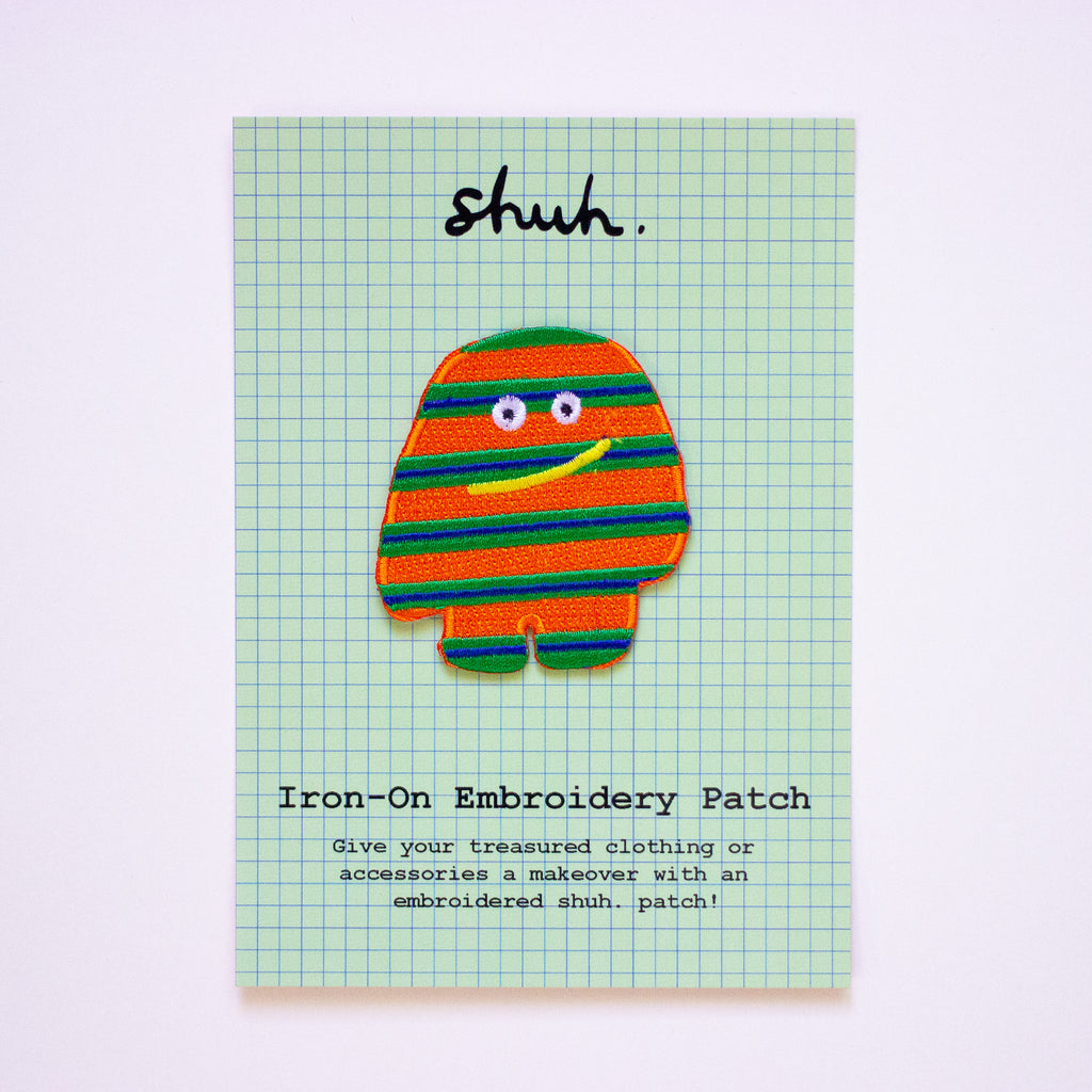 Striped character iron-on embroidery patch on a green grid card with 'shuh' text.