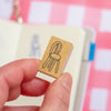 A hand holds a small wooden rubber stamp featuring a minimal black line drawing of a chair, with a blurred pink and white checkered notebook in the background.