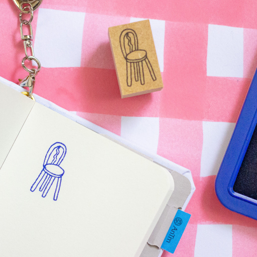 A wooden rubber stamp with a chair outline is shown above an open notebook that has the same chair stamped in blue ink. The stamp block is also visible standing upright.